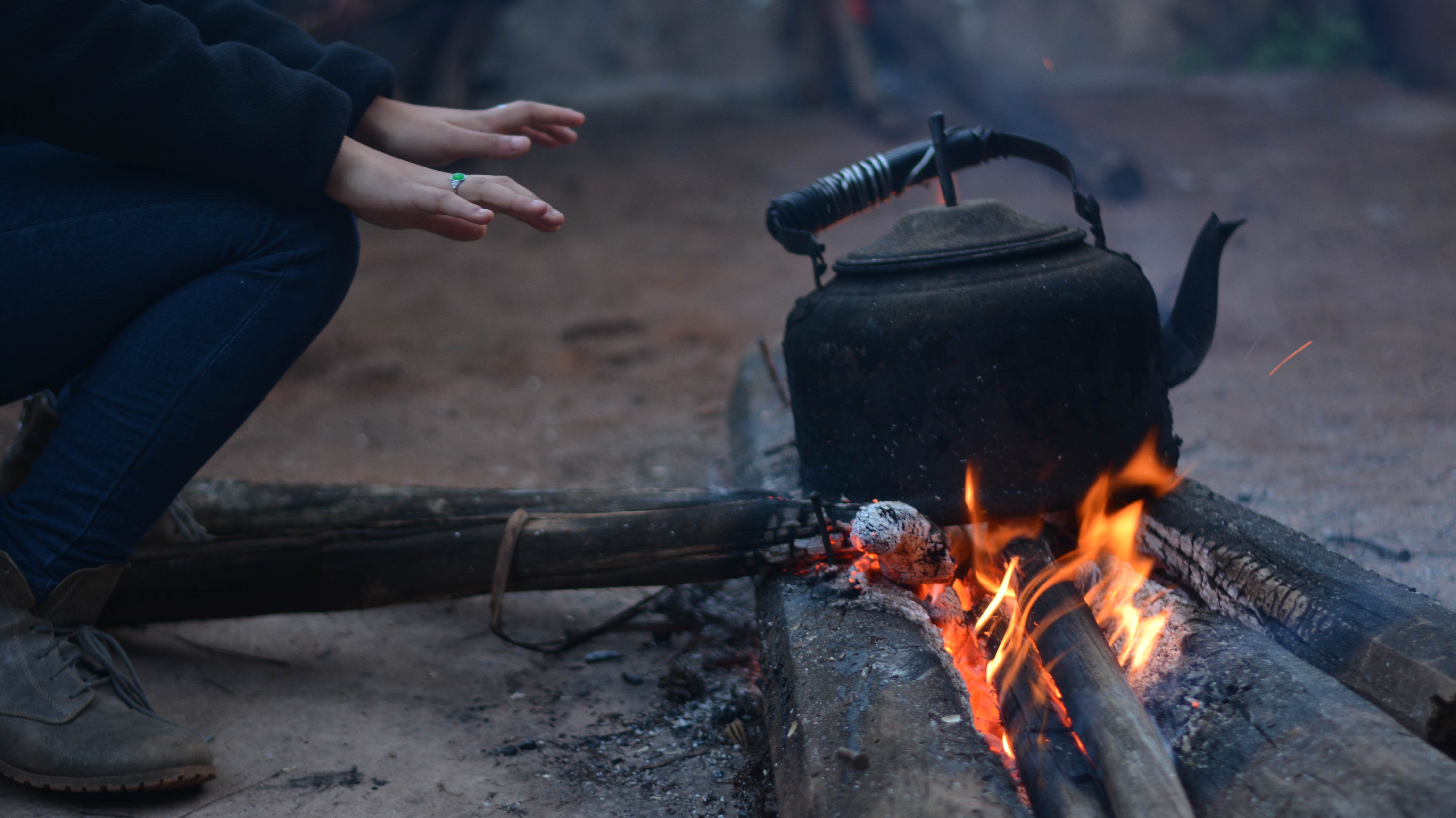 person warming their hands over a campfire with a kettle